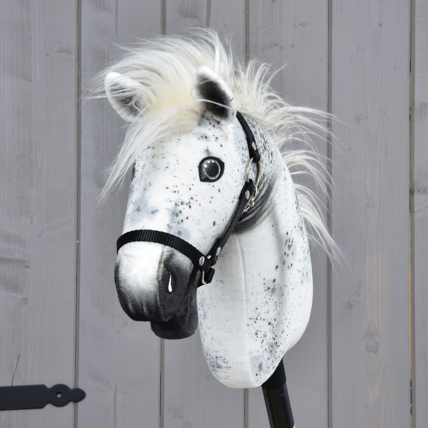 Hobby Horse Aspen s černou ohlávkou - velikost S od Hobby Horse LarDen, s bíločerným skvrnitým zbarvením a huňatou hřívou připomínající Appaloosu, nasazený na tyči a vystavený před pozadím z dřevěných panelů.