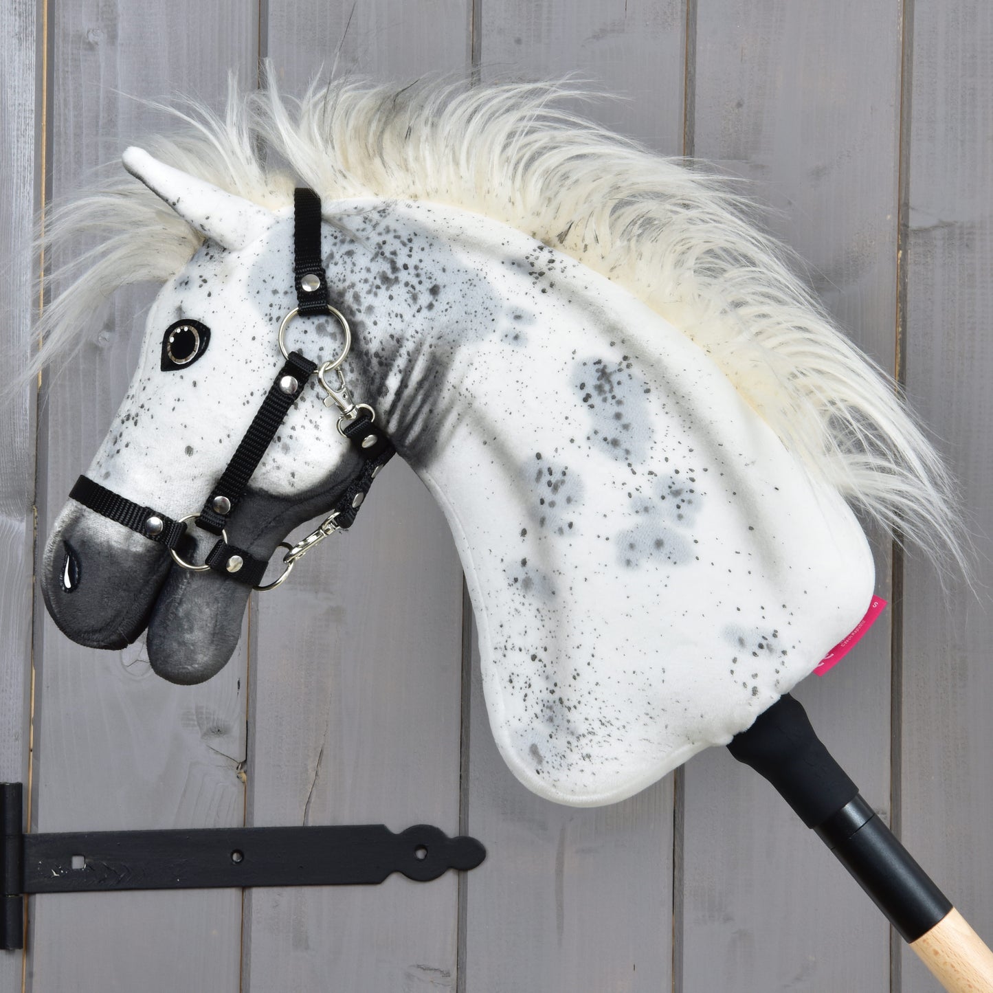 Hobby Horse LarDen "Hobby Horse Aspen with Black Halter", velikost S, s bílou plyšovou hlavou zdobenou černými skvrnami, huňatou hřívou a černou ohlávkou, nasazený na dřevěné tyči a vystavený před šedou dřevěnou stěnou.