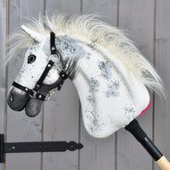 Hobby Horse LarDen "Hobby Horse Aspen with Black Halter", velikost S, s bílou plyšovou hlavou zdobenou černými skvrnami, huňatou hřívou a černou ohlávkou, nasazený na dřevěné tyči a vystavený před šedou dřevěnou stěnou.