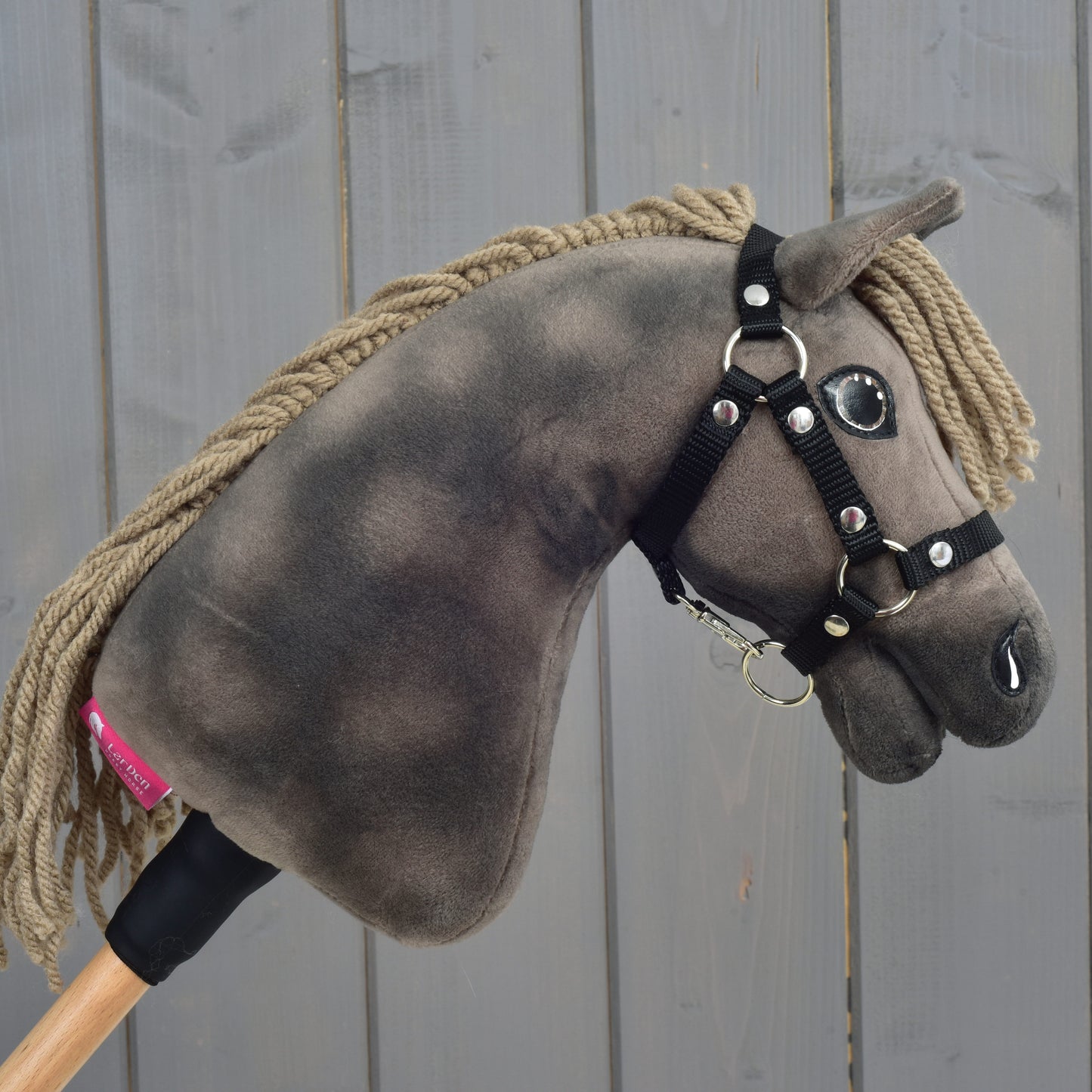 Hobby Horse Avalon s černou ohlávkou, velikost XS, od Hobby Horse LarDen, se šedočerným tělem, béžovou přízovou hřívou a na dřevěné tyči, zobrazený před šedým dřevěným plotem, ideální pro nápaditá parkurová dobrodružství.