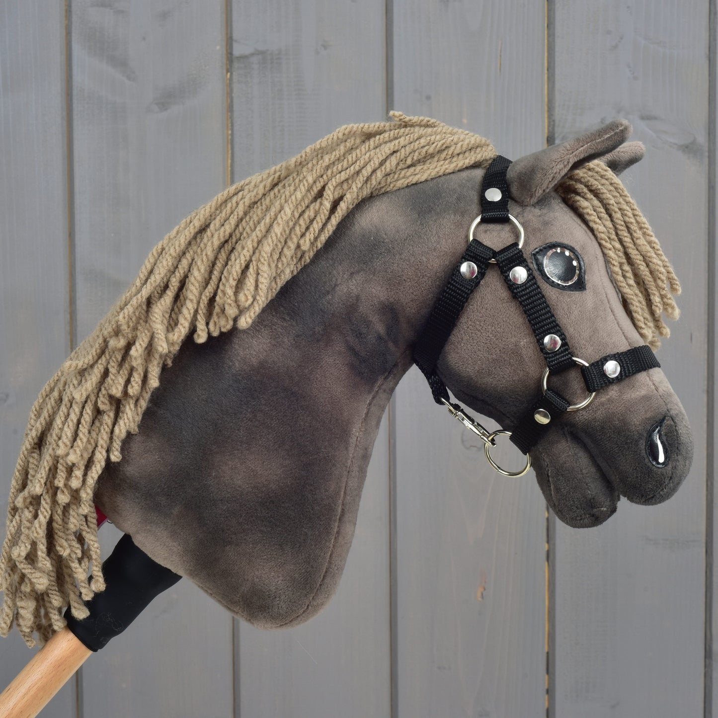 Hobby Horse Avalon s černou ohlávkou, velikost XS, od Hobby Horse LarDen, detail plyšového sportovního koně s hnědou přízovou hřívou, knoflíkovýma očima a černou uzdečkou na dřevěné tyči, na šedém dřevěném pozadí.