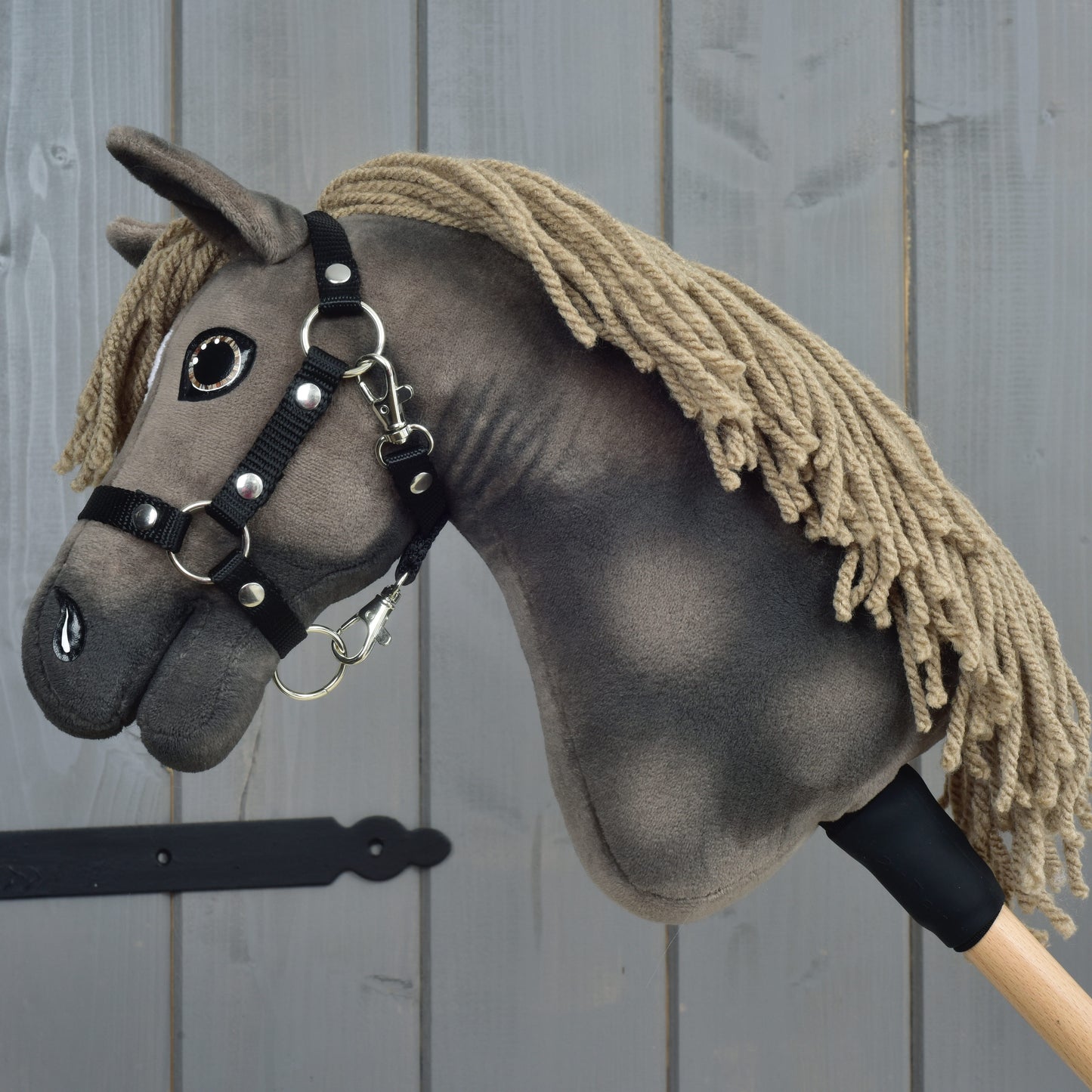 Hobby Horse Avalon s černou ohlávkou, velikost XS, od Hobby Horse LarDen, krásně ručně vyrobený Hobby Horse inspirovaný sportovním koněm plemene English Warmblood. Má béžovou přízovou hřívu, tmavě šedou látkovou hlavu a je nasazený na dřevěné tyči s elegantní černou ohlávkou. Vyfotografovaný před šedou dřevěnou stěnou, ideální pro nápaditou hru při parkurovém skákání.