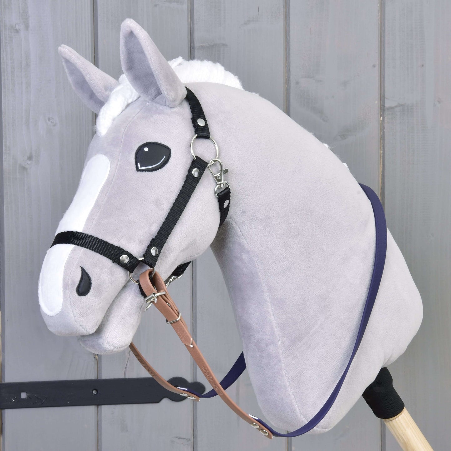 Plyšový Hobby Horse od Hobby Horse LarDen se světle šedou hlavou, bílou hřívou a znaky na obličeji má nasazené Otěže pogumované Brown Purple. Je umístěný před dřevěnou stěnou.