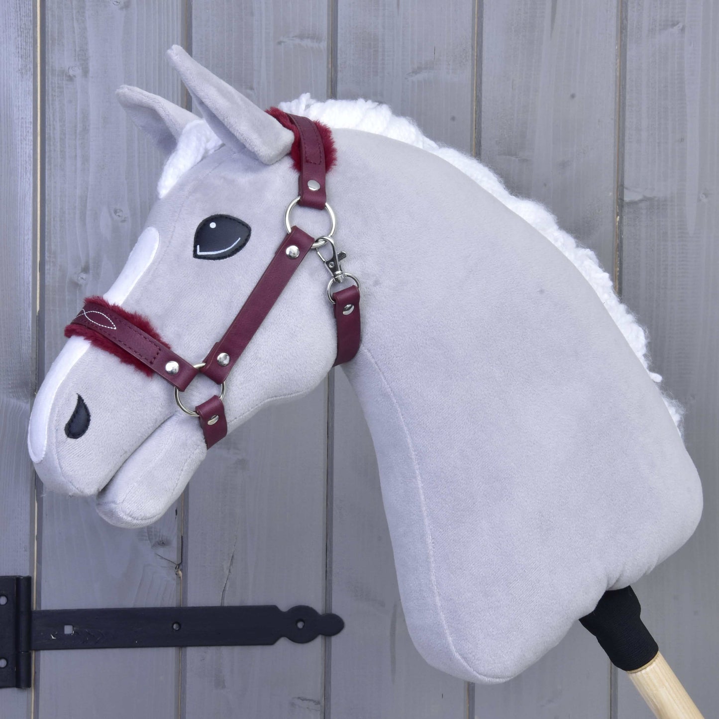 Ohlávka s beránkem z koženky Burgundy od Hobby Horse LarDen na šedém plyšovém Hobby Horse s bílou hřívou a vínovou uzdečkou dodává stylový vzhled na pozadí šedé dřevěné stěny — je tak dokonalým doplňkem.
