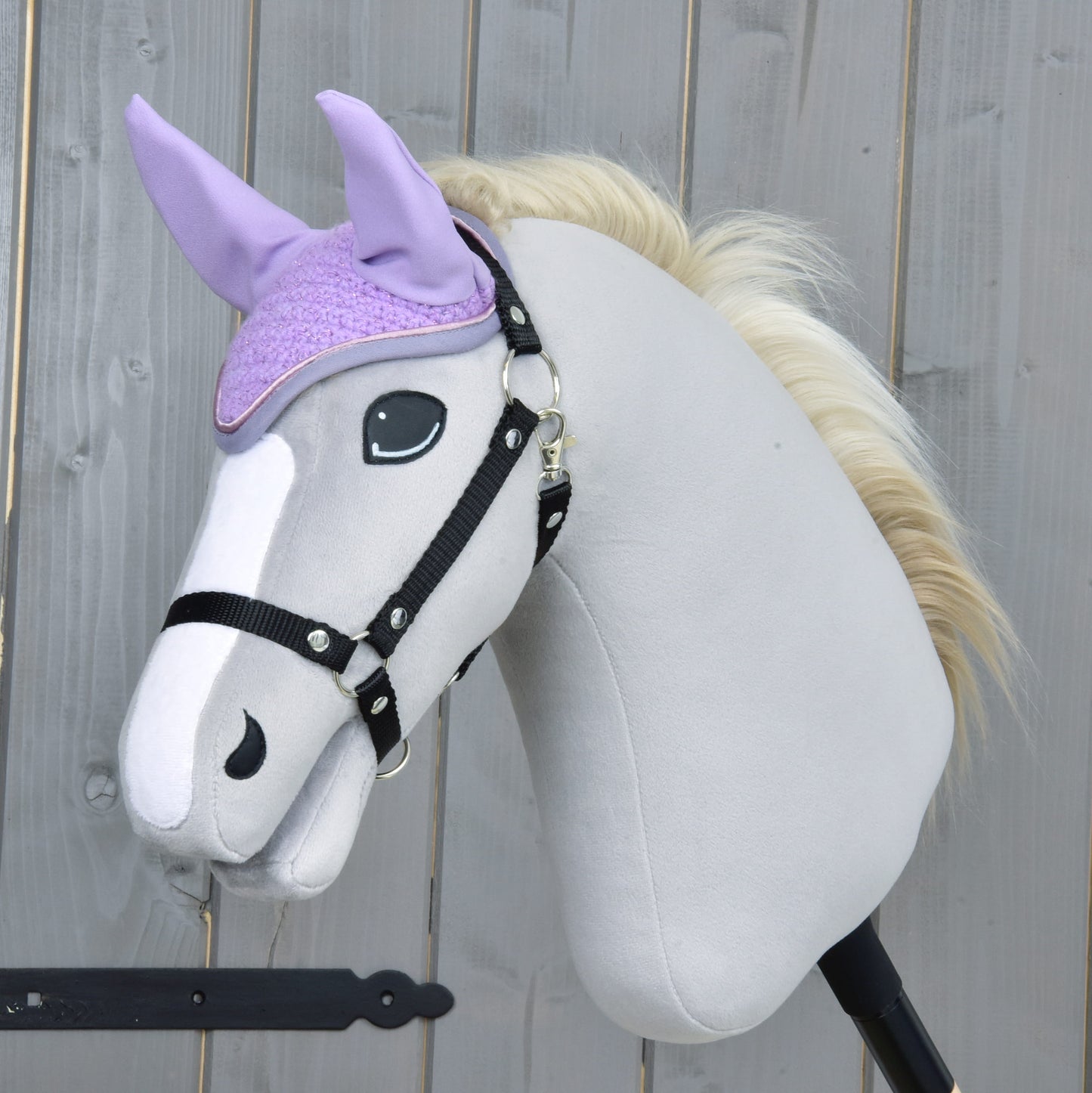 Plyšový Hobby Horse se světle šedým tělem, černou uzdečkou a krémovou hřívou nosí Čabraka Orchid od Hobby Horse LarDen pro ochranu před hmyzem. V pozadí je dřevěný plot.