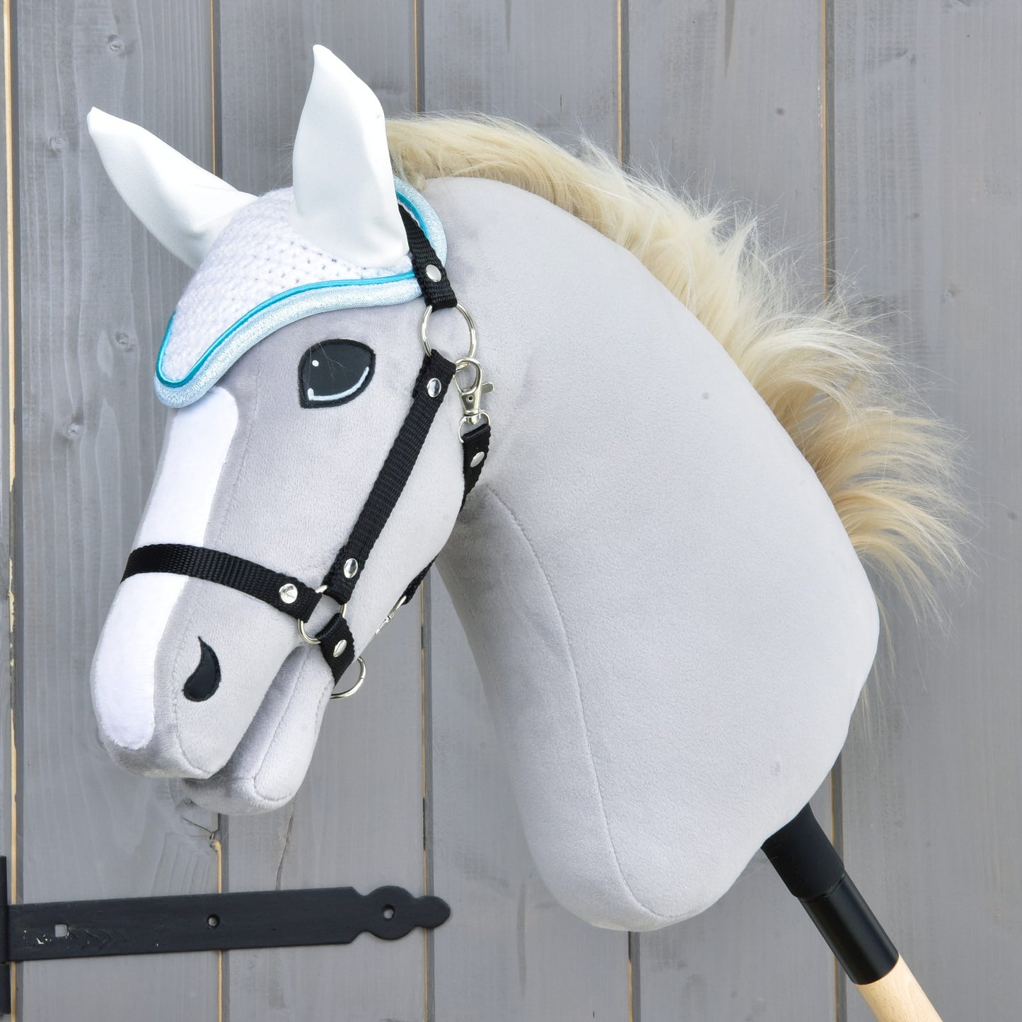 Detail šedého plyšového Hobby Horse od Hobby Horse LarDen s bílou hřívou, černou uzdečkou a čabrakou háčkovanou Glitter Cuties pro ochranu proti mouchám, nasazeného na dřevěné tyči před dřevěným plotem.