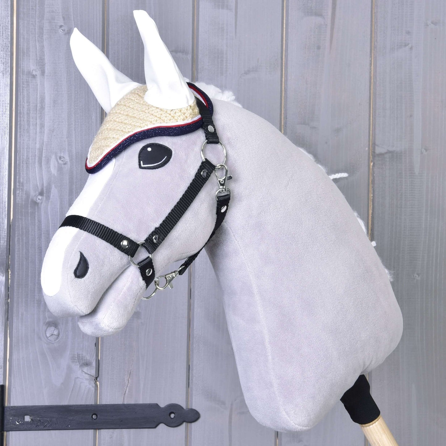 Šedý plyšový Hobby Horse od Hobby Horse LarDen s červenou háčkovanou čabrakou Nicol, uzdečkou a slaměným kloboukem, nasazený na dřevěné tyči a zobrazený před pozadím z dřevěných prken.