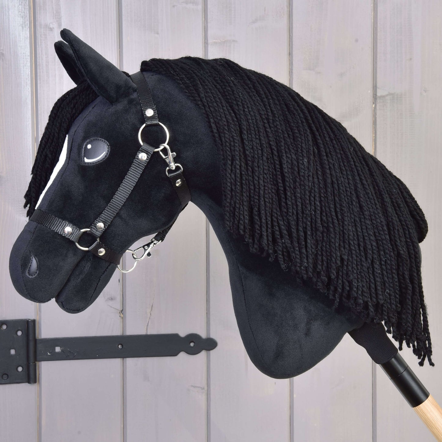 Hobby Horse Cloony s černou ohlávkou od Hobby Horse LarDen, černý hřebec s bílou skvrnou na hlavě, černou přízovou hřívou a uzdečkou na dřevěné tyči, před světle šedými dřevěnými prkny.