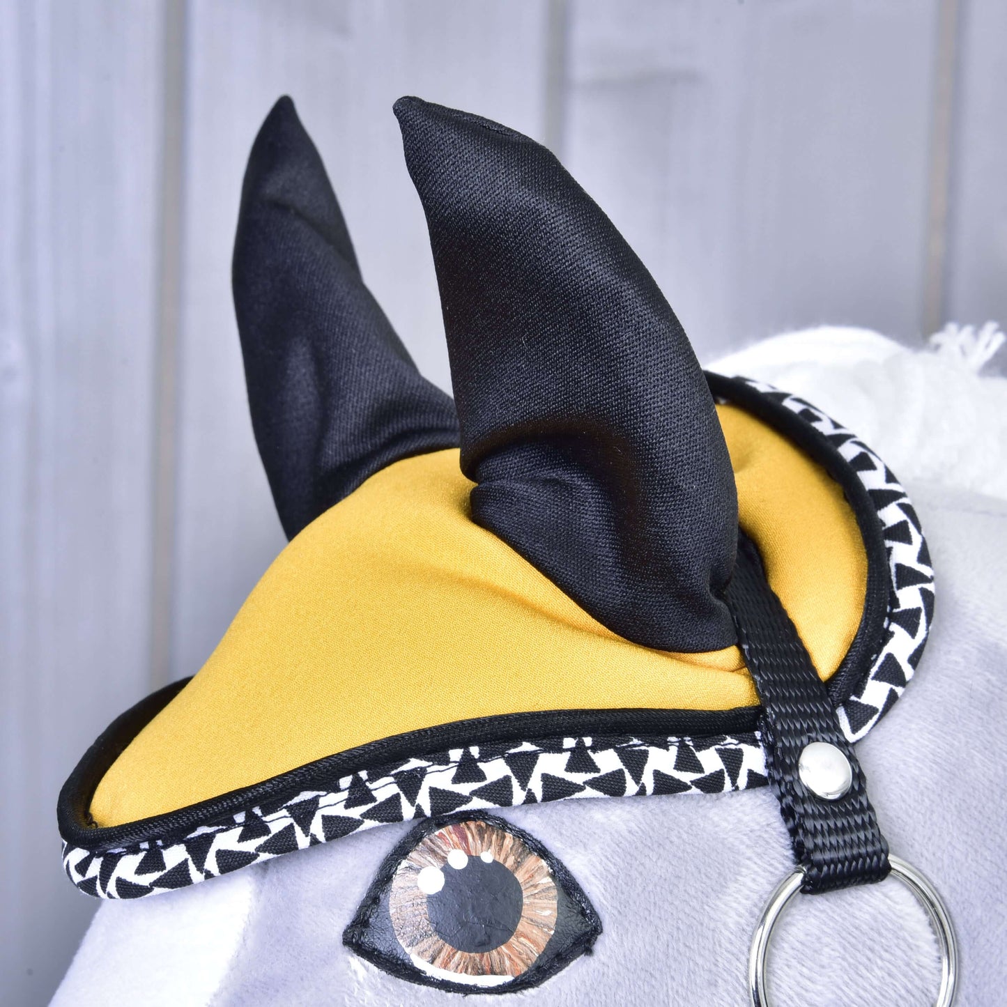 Detail Set Yellow Beauty od Hobby Horse LarDen ukazuje jeho plyšovou hlavu se žluto-černým ušním návlekem, geometrickým černobílým lemováním, malovaným okem a obličejem s bílou srstí na pozadí šedého dřeva.