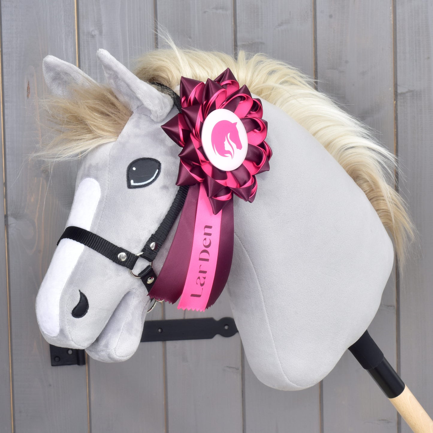 Plyšový Hobby Horse Kokarda Raspberry Rose od Hobby Horse LarDen se šedým obličejem, krémovou hřívou a černou uzdečkou na tyči, ozdobený velkou růžovo-bílou drezurní kokardou s logem LarDen na krku. Pozadí tvoří dřevěné obložení.
