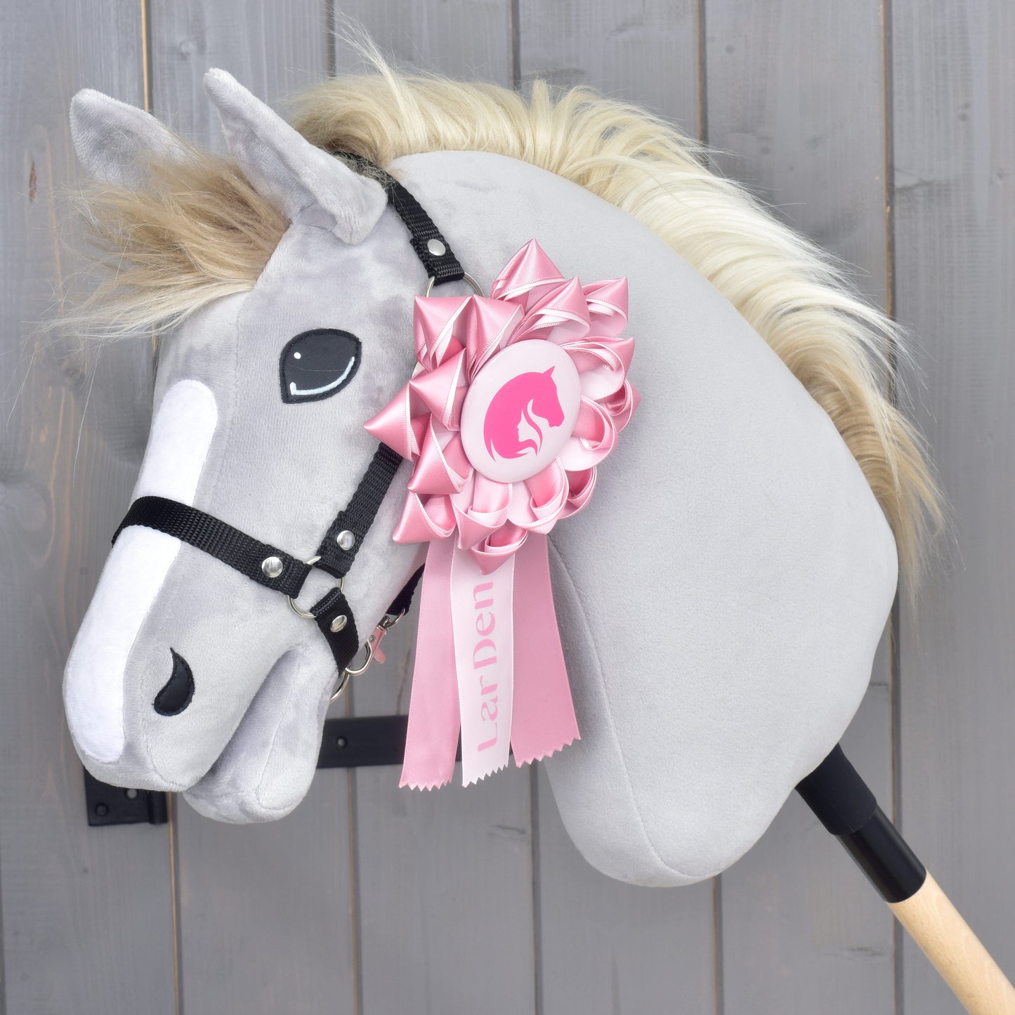 Hobby Horse LarDen „Hobby Horse Kokarda LarDen Pink“, šedý plyšový kůň s černou uzdečkou a krémovou hřívou, ozdobený velkou růžovou výstavní kokardou na hlavě, na dřevěném pozadí.