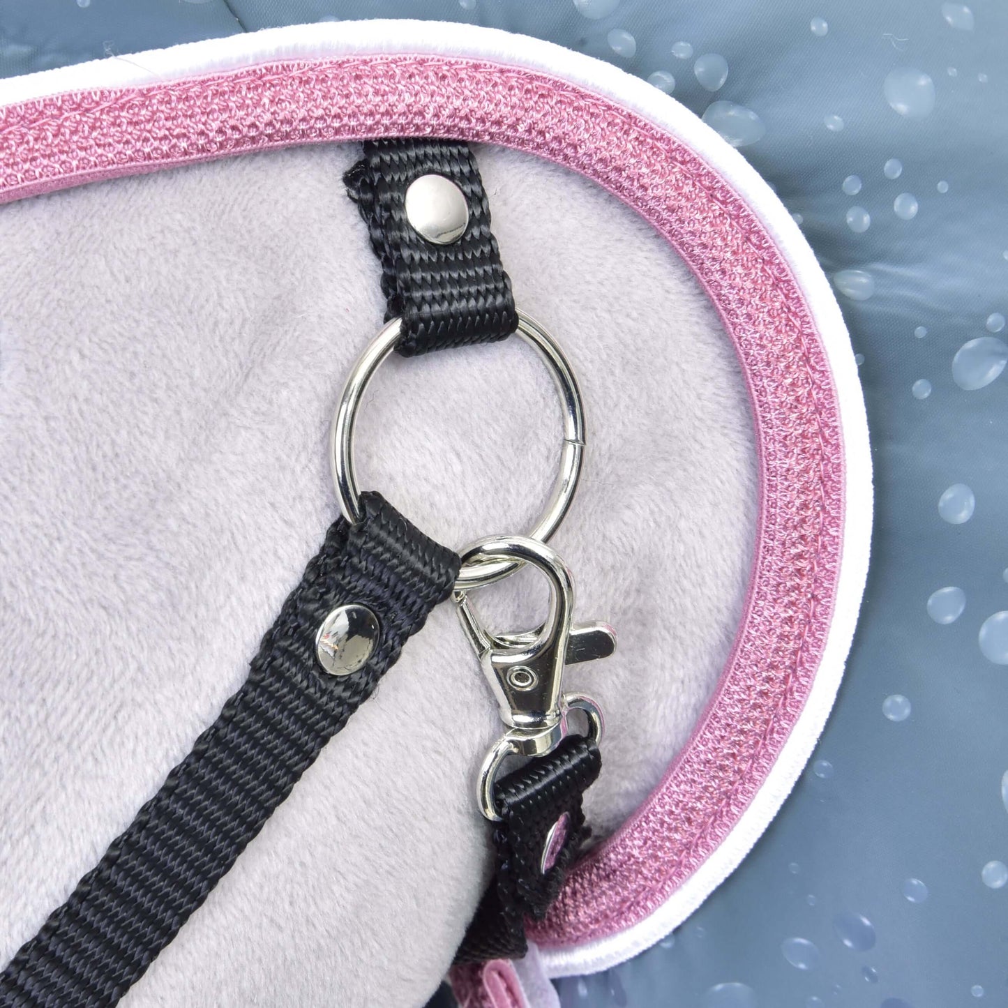 Detail černého popruhu s kovovou sponou a kroužkem připevněného k Raincoat Best od Hobby Horse LarDen se světle šedou látkou a růžovým lemováním. Modré pozadí s kapkami vody zdůrazňuje odolnost do každého počasí pro Hobby Horse zábavu.