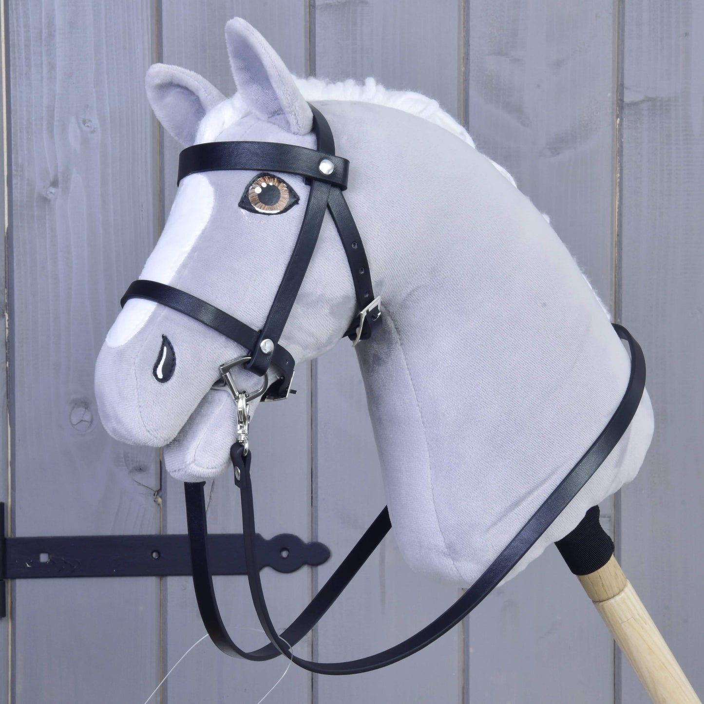 Hobby Horse LarDen Set Basic, šedý plyšový Hobby Horse s černou uzdečkou a otěžemi na dřevěné tyči, je zobrazen před šedými dřevěnými panely.