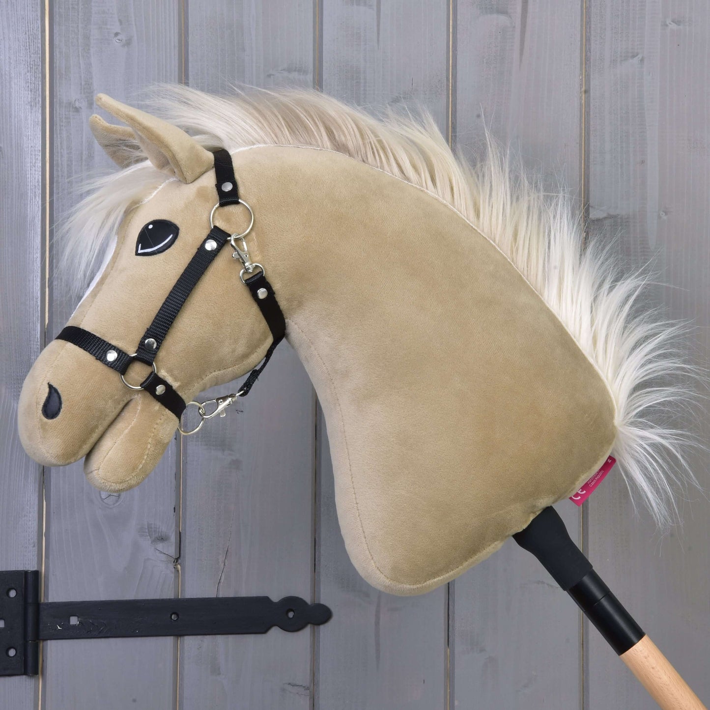 Hobby Horse Dancing Lady s černou ohlávkou od Hobby Horse LarDen s plyšovou hlavou klisny anglického plnokrevníka v béžové barvě a s bílou hřívou na dřevěné tyči, zobrazený před šedou stěnou z dřevěných panelů.