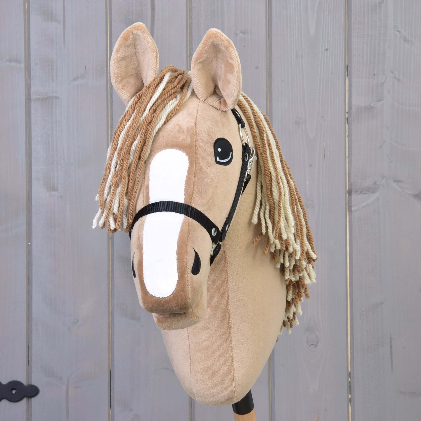 Hobby Horse Bren s černou ohlávkou od Hobby Horse LarDen, se světle hnědým plyšovým tělem, bílou lysinou a přízovou hřívou v krémové a hnědé barvě, před světlým dřevěným pozadím.