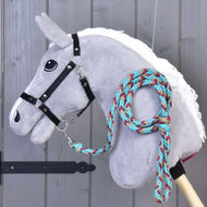 Hobby Horse - pletené vodítko tyrkys od Hobby Horse LarDen na šedé plyšové hlavě na dřevěné tyči s černou uzdečkou a pleteným vodítkem v tyrkysové, červené a béžové barvě omotaným kolem krku, před dřevěným obkladem.