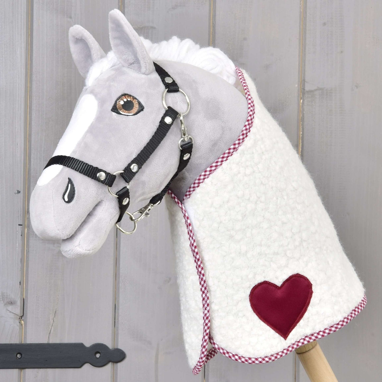 Plyšový Hobby Horse od Hobby Horse LarDen s bílošedým obličejem, černou uzdečkou a fleecovou Dekou Cube Red s červenou nášivkou ve tvaru srdce, nasazený na dřevěné tyči na pozadí světlého dřeva.