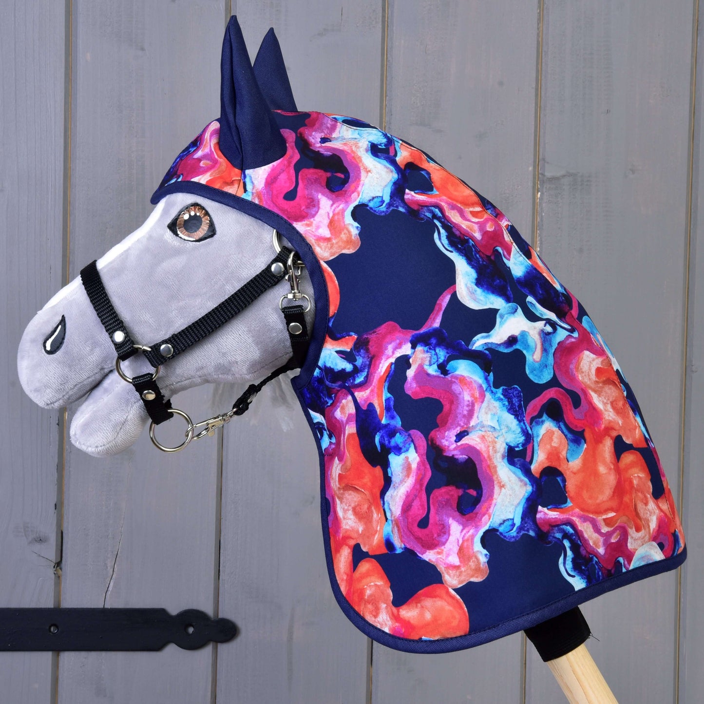 Pláštěnka Rainbow (zimní softshell) od Hobby Horse LarDen na plyšovém Hobby Horse s černou ohlávkou a výrazným zimním softshellovým krytem hřívy a krku v modrých, růžových, oranžových a fialových vzorech, zobrazeném před šedým dřevěným pozadím.