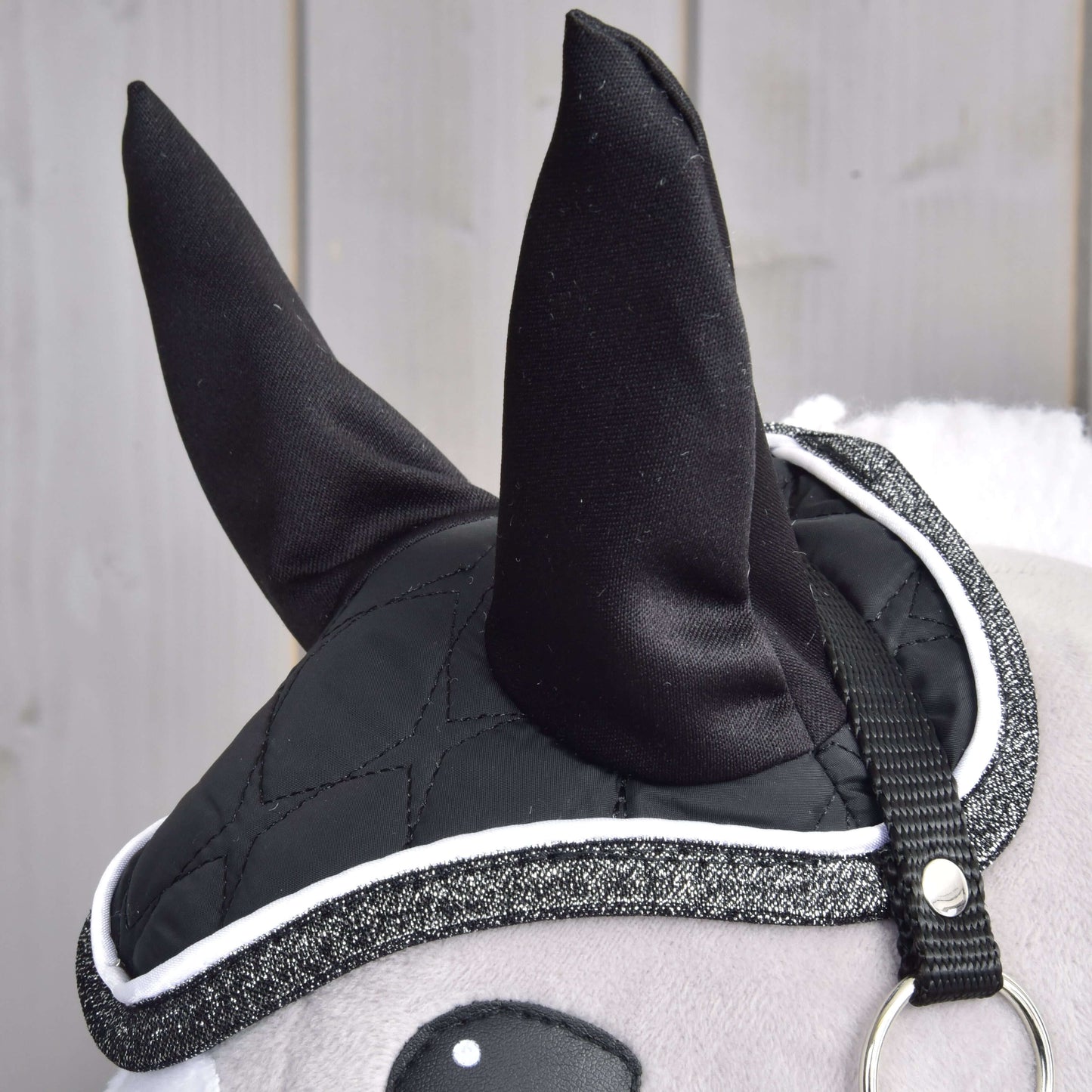 Detail Set Thermo Black Queen od Hobby Horse LarDen s plyšovými černými ušima s čabrakou, prošívaným krytím s třpytivým lemem jako deka pro Hobby Horse a páskem s kovovým kroužkem, před světlým dřevěným obložením.