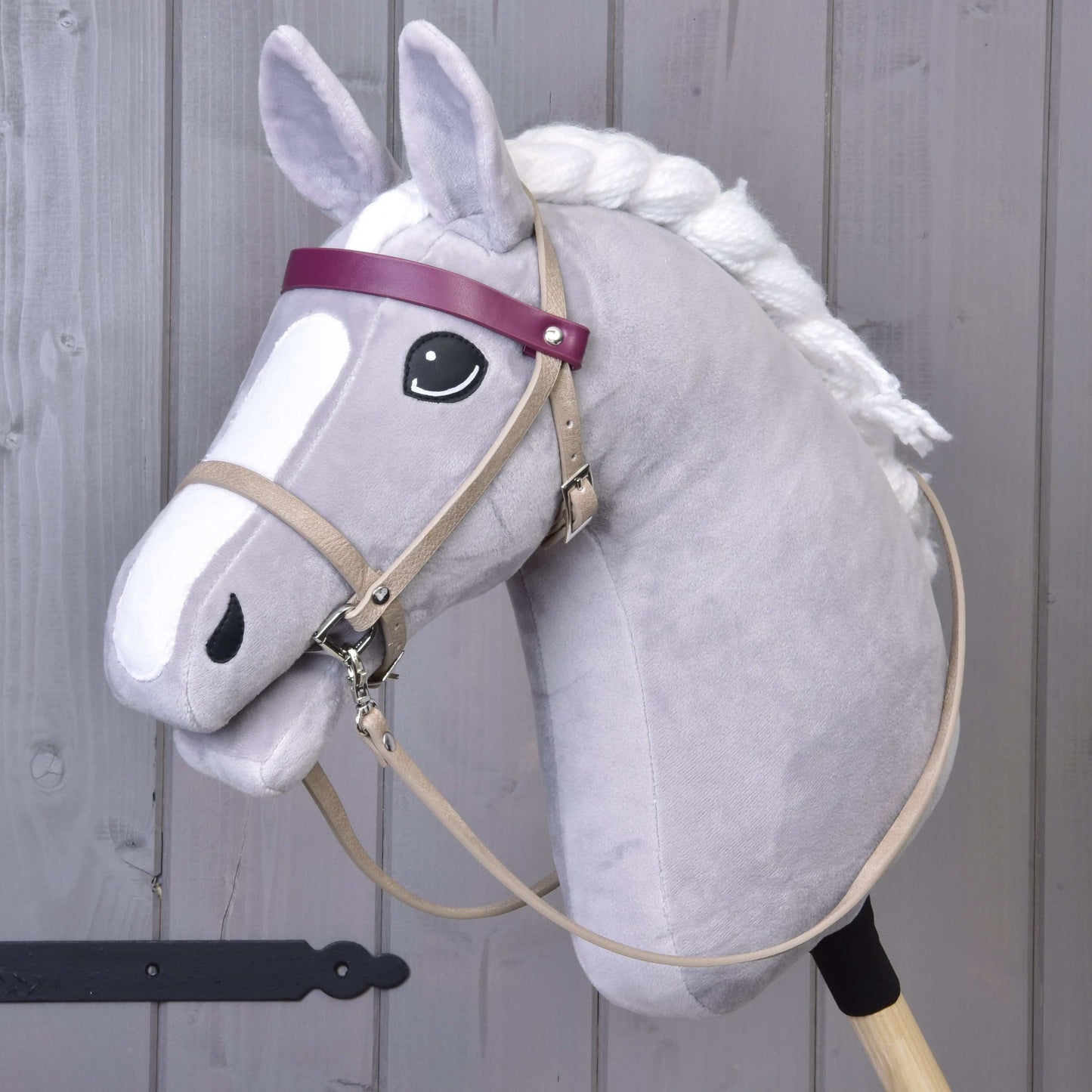 Hobby Horse LarDen se šedým Hobby Horse s bílou hřívou, černým okem a koženkovým setem Bordeaux – uzdečka, otěže a martingal –, doplněným o fialovou čelenku a obličej v kresleném stylu. Stojí vzpřímeně před dřevěným pozadím.