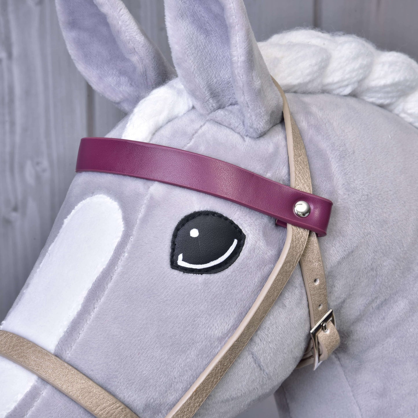 Detail Hobby Horse LarDen s bílou hřívou a vyšitým černým okem, který má na sobě koženkový set Bordeaux – uzdečka, otěže a martingal. Má fialovou čelenku a je vyobrazený před dřevěným pozadím.