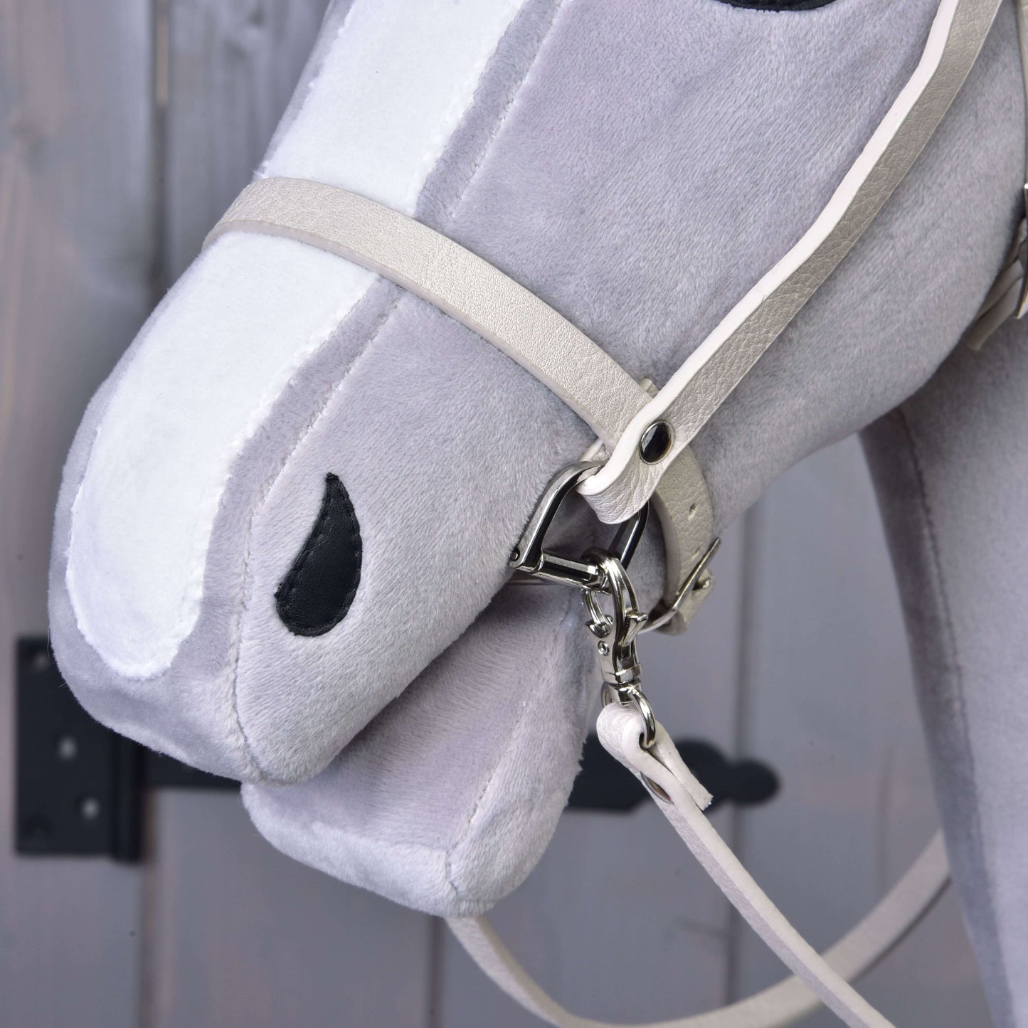 Detail hlavy šedo-bílého Hobby Horse s koženkovým setem Aqua – uzdečka, otěže a martingal – od Hobby Horse LarDen. Uzdečka má kovové kování. Rozmazané pozadí ukazuje dřevěný povrch a tmavý kovový pant.