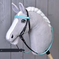 Šedý plyšový Hobby Horse s bílou hřívou a bílými znaky na hlavě má Kompletní uzdečku s tyrkysovou čelenkou bez nánosníku od Hobby Horse LarDen, černé otěže a je upevněný na dřevěné tyči před šedou dřevěnou stěnou.