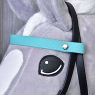 Detail hlavy šedého plyšového koně Hobby Horse LarDen s Kompletní uzdečkou s tyrkysovou čelenkou bez nánosníku. Součástí jsou černá otěž, bílé ucho, bílé znaky na hlavě a oko v kresleném stylu s černým obrysem a bílým odleskem.