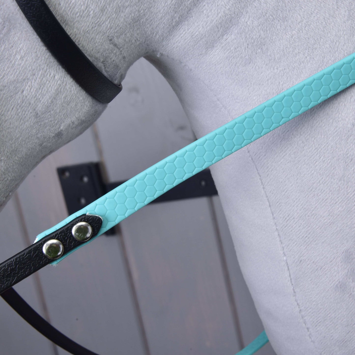Detail Kompletní uzdečky s tyrkysovou čelenkou bez nánosníku od Hobby Horse LarDen s texturovaným tyrkysovým řemínkem z koženky ve vzoru včelích pláství s kovovými nýty, připevněným k černému řemínku na světle šedém povrchu s rozmazaným dřevěným pozadím.