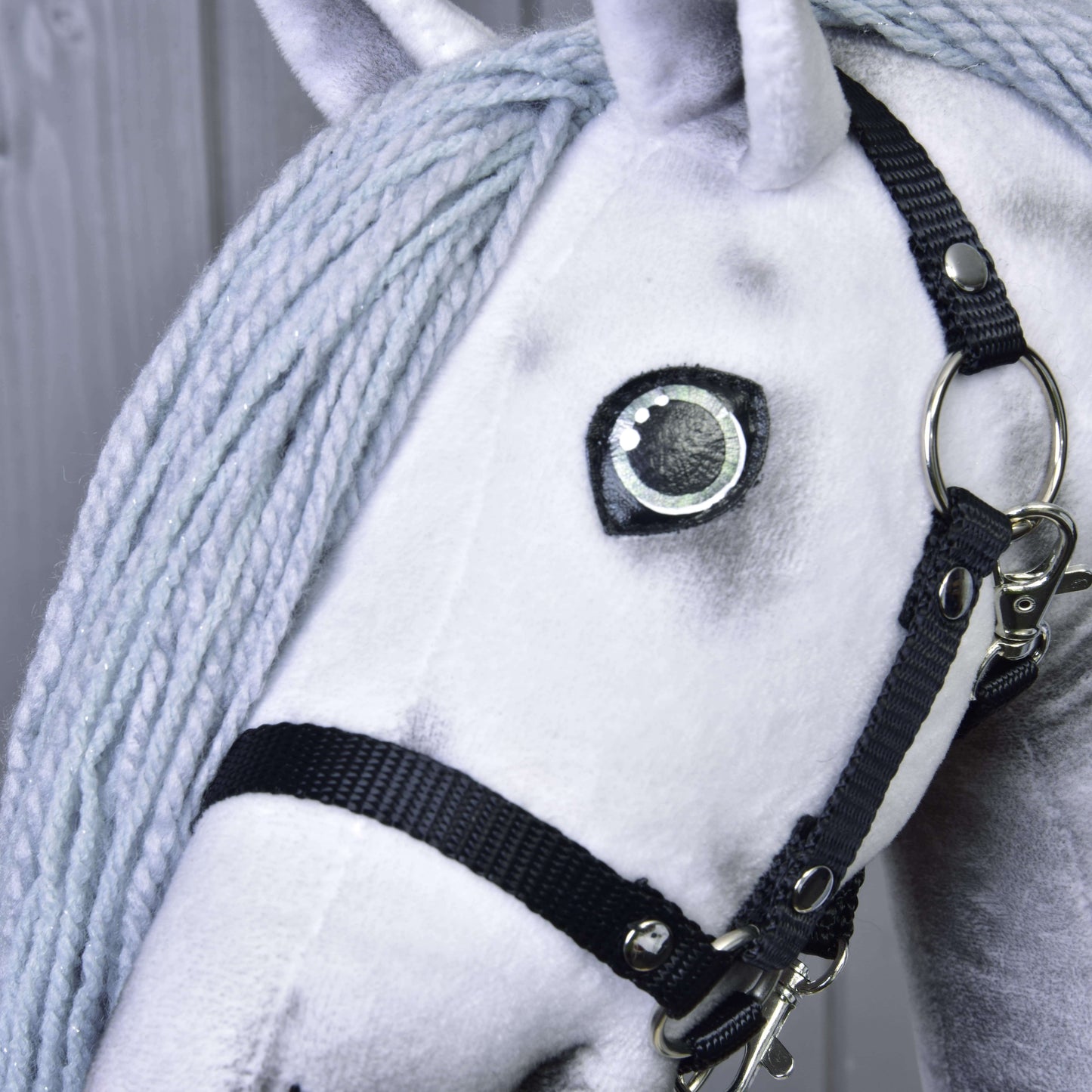 Detail Hobby Horse jednorožec Silver s uzdou a ohlávkou od Hobby Horse LarDen se silnou světle šedou hřívou z příze, černou uzdou a velkým šitým okem. Vyniká jeho plyšová textura, zatímco v pozadí je rozmazaný dřevěný plot.