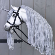 Hobby Horse jednorožec Silver s uzdou a ohlávkou od Hobby Horse LarDen s bílým obličejem, šedou hřívou z příze, světle šedým rohem a černou uzdou, nasazený na tyči a vyfotografovaný na dřevěném pozadí.