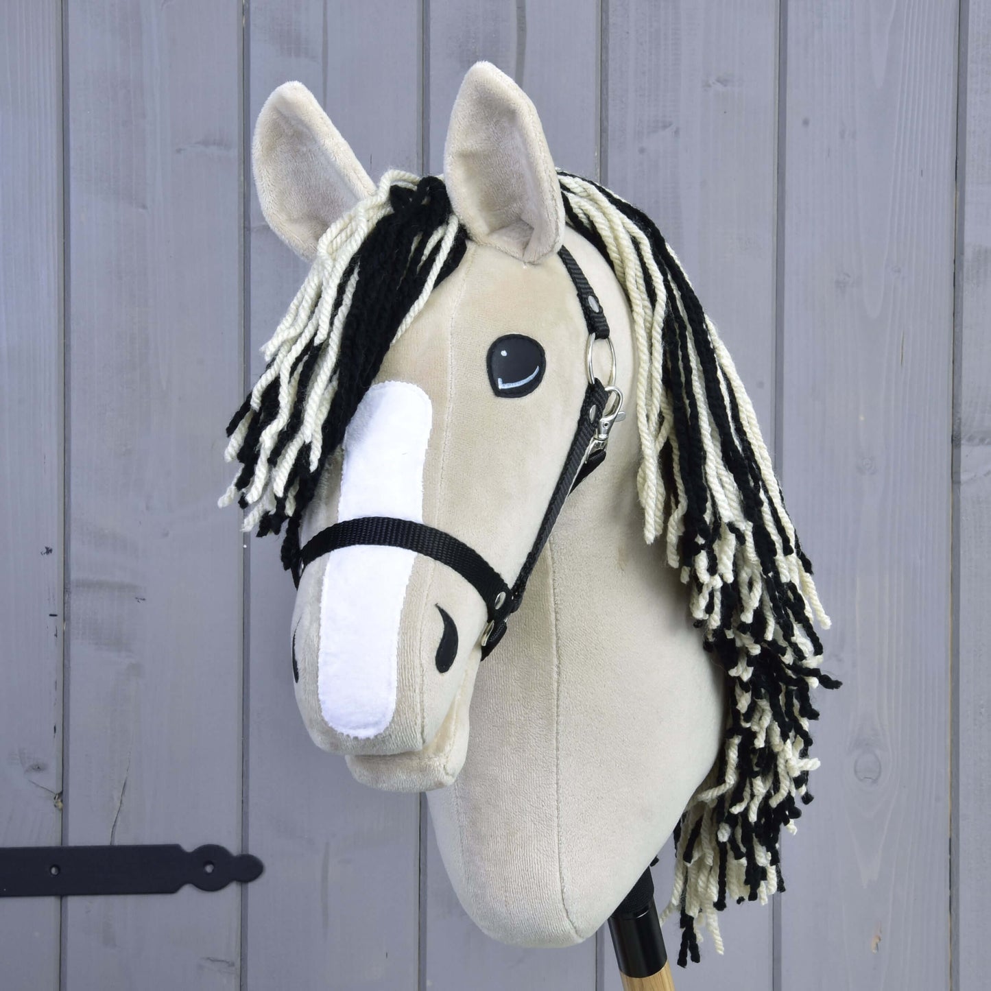 Hobby Horse Aurora s černou ohlávkou od Hobby Horse LarDen, s béžovým plyšovým provedením inspirovaným American Quarter Horse, černou ohlávkou, černo-bílou přízovou hřívou, bílým plstěným pruhem na nose a na dřevěné tyči před světle šedou dřevěnou stěnou.