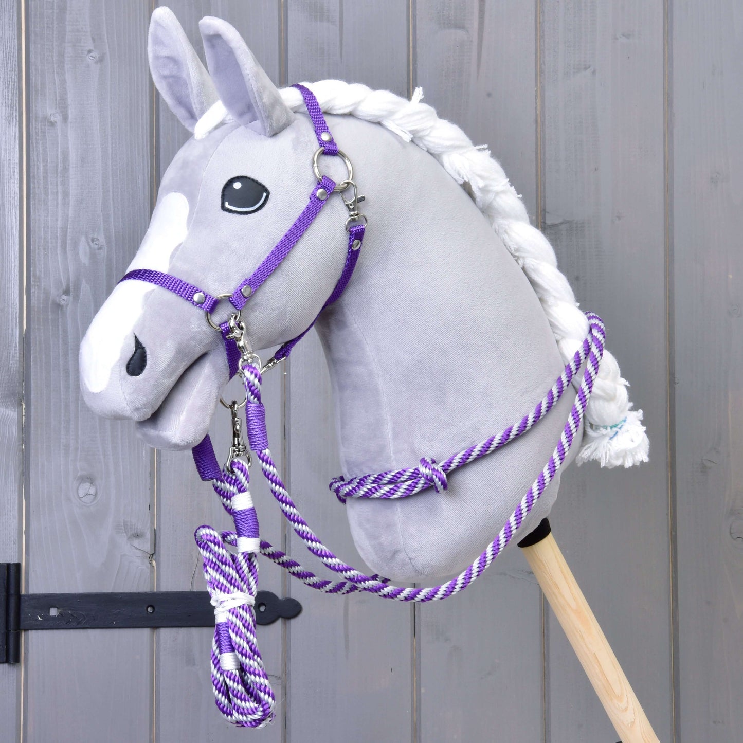 Plyšový Hobby Horse se šedou hlavou, bílou hřívou a bílou lysinou má sadu Set Start Fialový (ohlávka + nákrčník + vodítko + otěže) od Hobby Horse LarDen a je upevněný na dřevěné tyči před šedou dřevěnou stěnou.