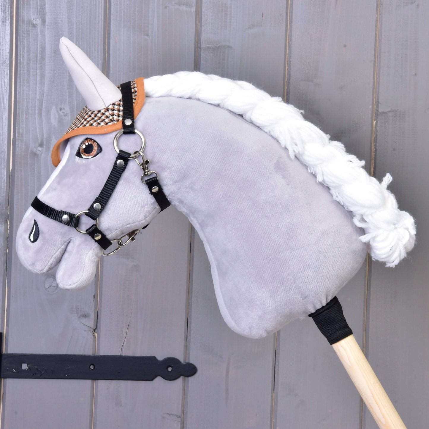 Čabraka anglická kostka hnědá od Hobby Horse LarDen na plyšovém Hobby Horse se šedou hlavou, černou uzdečkou, hnědou kostkovanou čabrakou na ochranu proti hmyzu a bílou hřívou, nasazeném na dřevěné tyči před dřevěným obkladem.