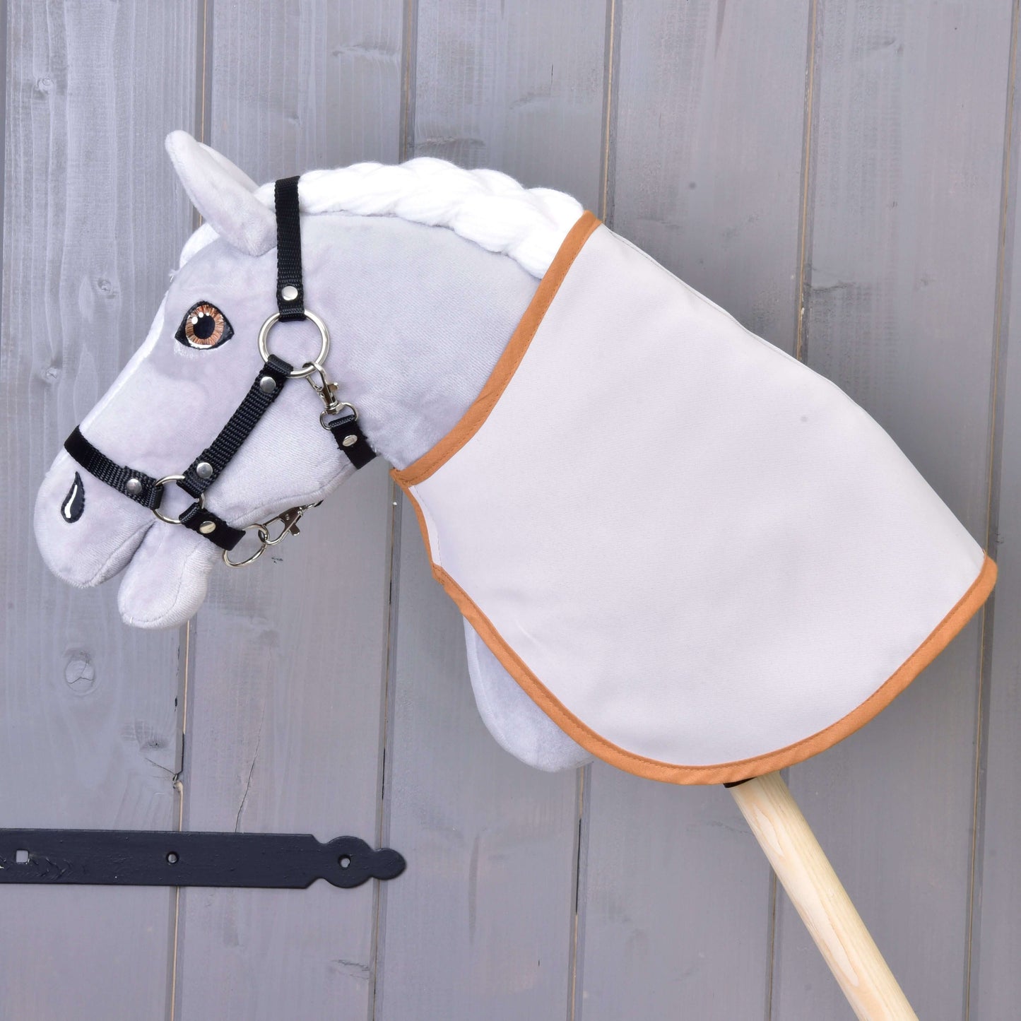 Šedý Hobby Horse LarDen s bílou hřívou, černou uzdečkou a Dekou anglická kostka, hnědá a oboustranná, nasazený na dřevěné tyči před šedou dřevěnou stěnou.