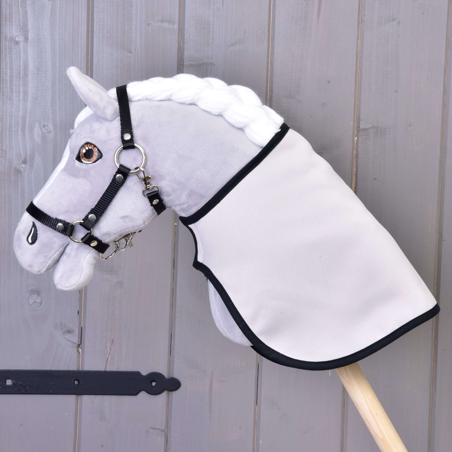 Šedý plyšový Hobby Horse s bílou hřívou, černou uzdečkou a Dekou anglická kostka černá oboustranná od Hobby Horse LarDen, nasazený na dřevěné tyči na pozadí stěny z dřevěných prken.