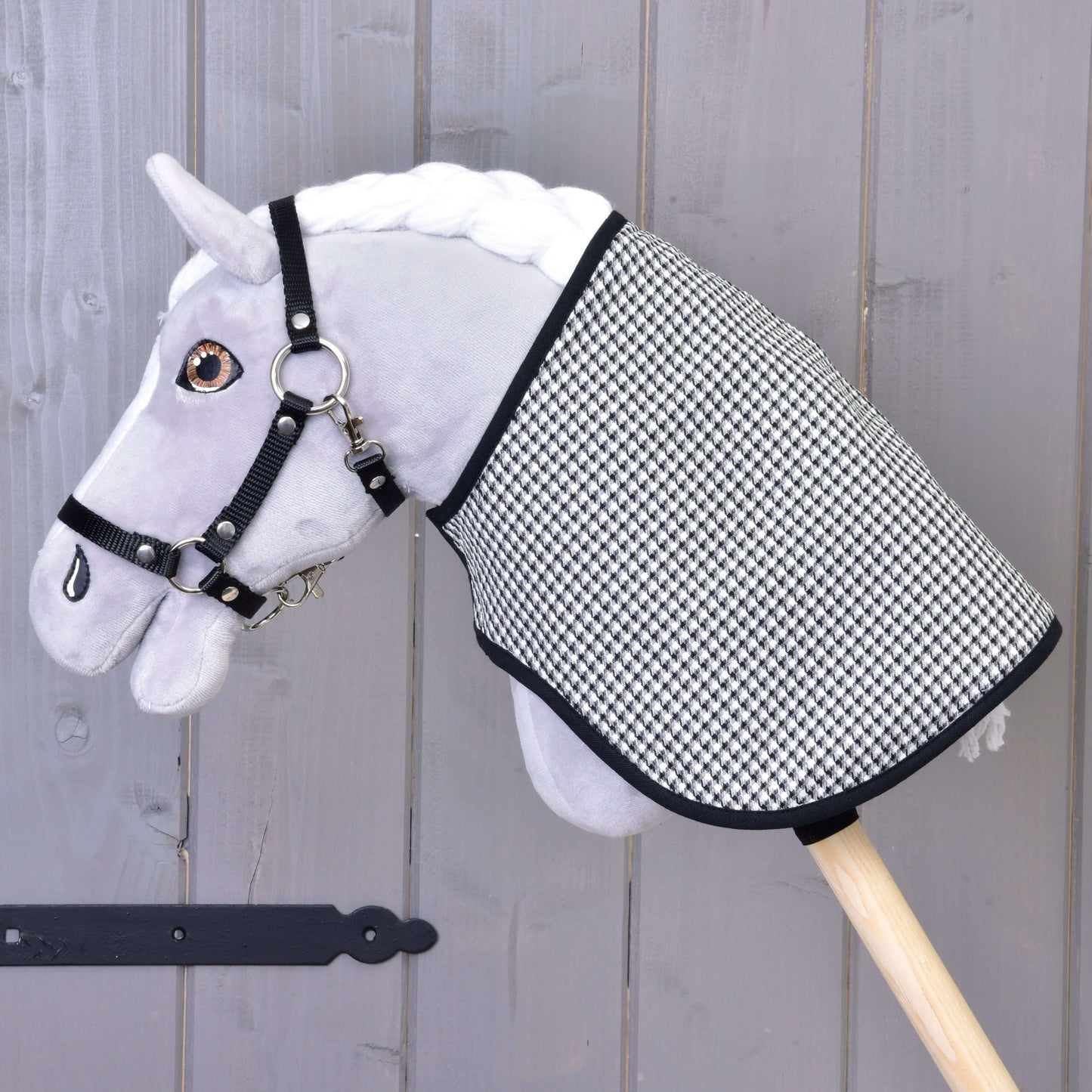 Šedý plyšový Hobby Horse s černou uzdečkou a Dekou anglická kostka černá oboustranná od Hobby Horse LarDen, nasazený na dřevěné tyči na pozadí šedé dřevěné stěny.