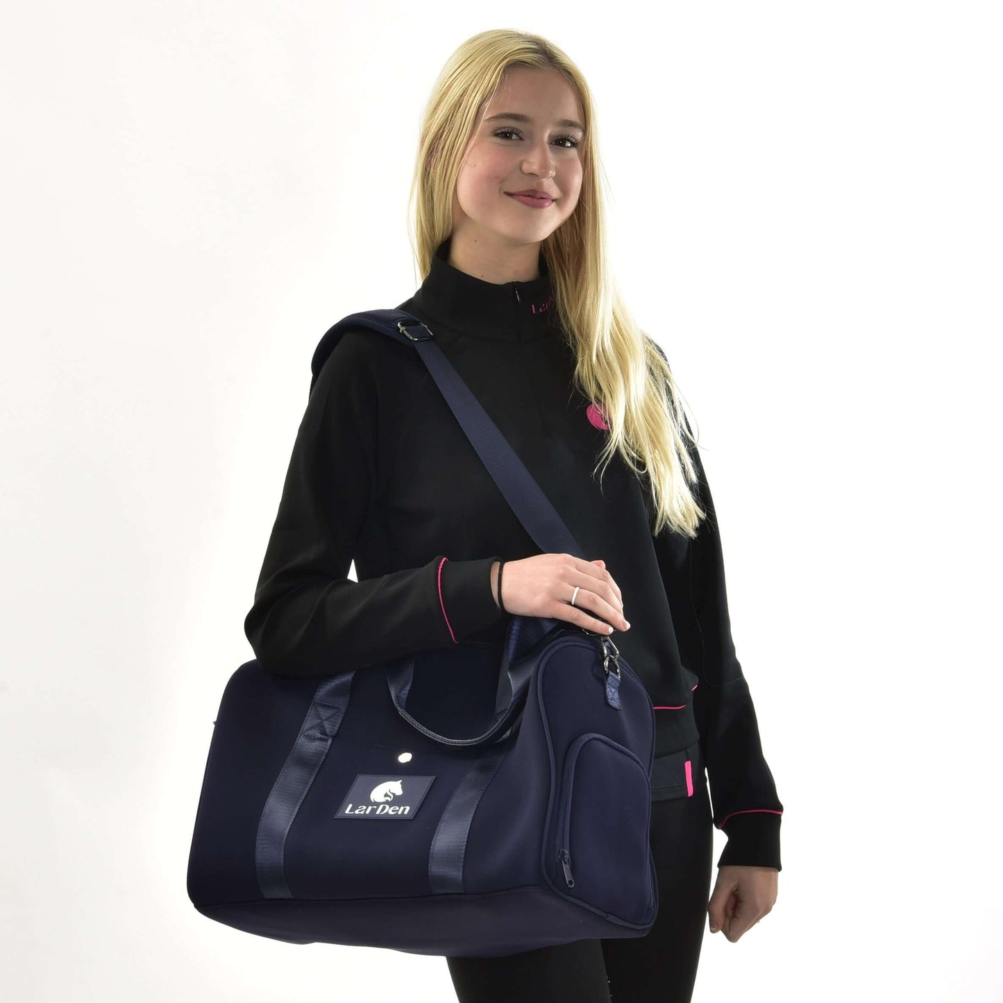 Usměvavá žena s dlouhými blond vlasy v černé bundě s růžovým lemováním stojí na bílém pozadí a drží Sportovní tašku LarDen od Hobby Horse LarDen, tmavě modrou tašku s nastavitelným popruhem, ideální pro každodenní použití.