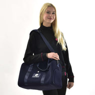 Usměvavá žena s dlouhými blond vlasy v černé bundě s růžovým lemováním stojí na bílém pozadí a drží Sportovní tašku LarDen od Hobby Horse LarDen, tmavě modrou tašku s nastavitelným popruhem, ideální pro každodenní použití.