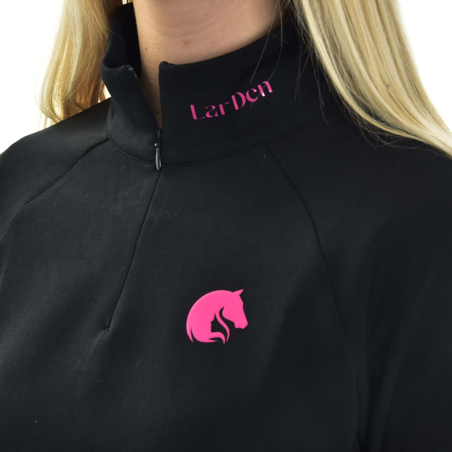 Osoba s dlouhými blond vlasy má na sobě mikinu LarDen od Hobby Horse LarDen – černý top na zip s růžovým logem koňské hlavy na hrudi a nápisem Lar Den v růžové barvě u límce.