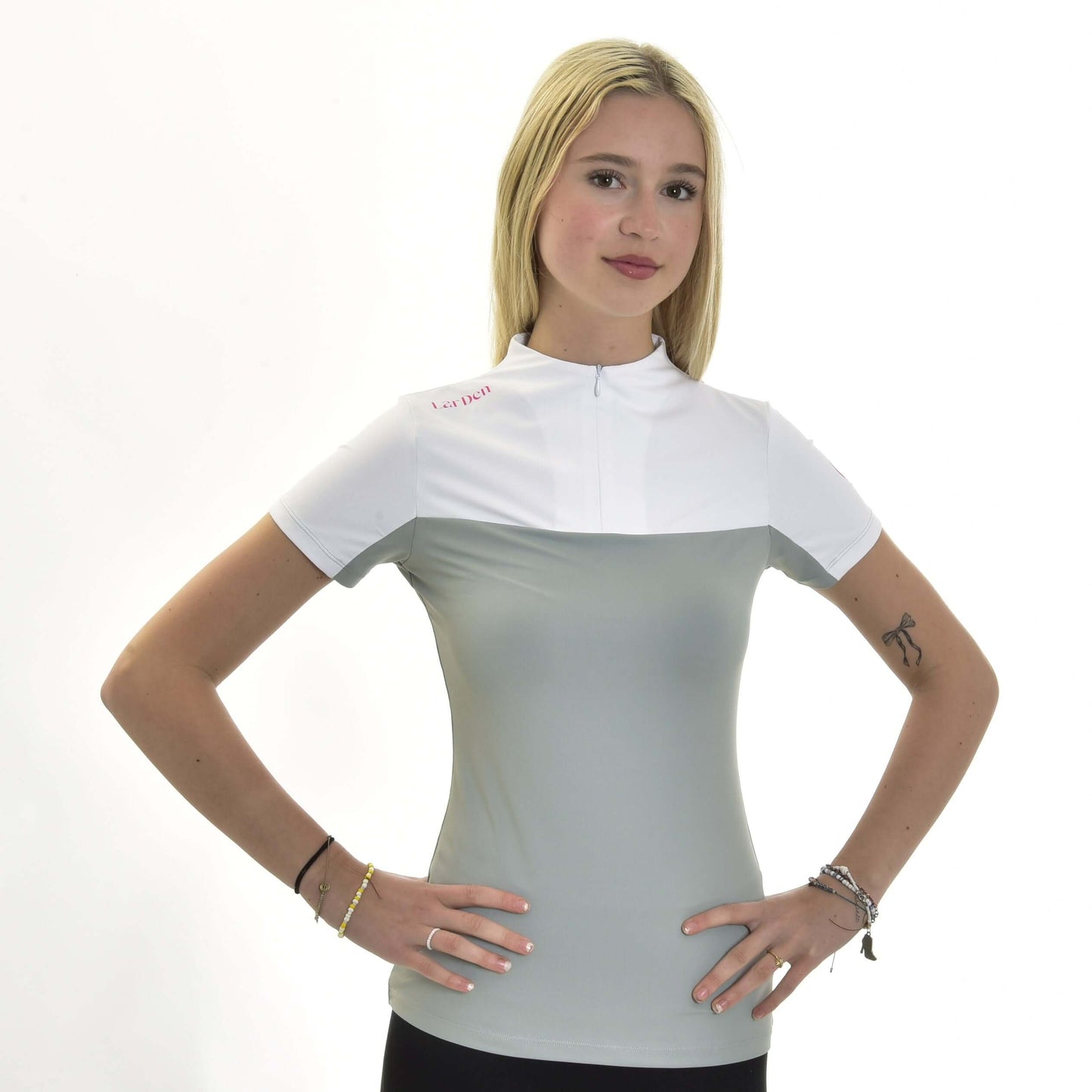 Mladá žena s dlouhými blond vlasy má na sobě Sportovní triko LarDen Premium od Hobby Horse LarDen, sportovní tričko s krátkým rukávem ze světle šedého a bílého spandexu. Stojí čelem ke kameře s rukama v bok a jsou vidět náramky a malé tetování na levé paži.