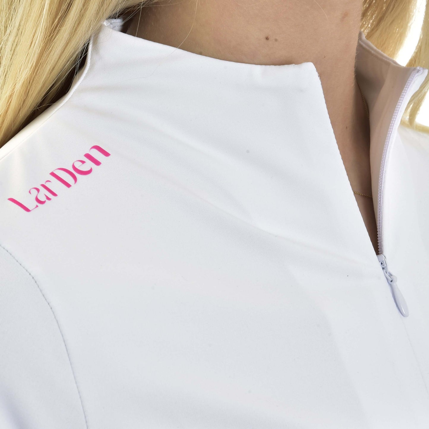 Detail blond osoby ve Sportovním triku LarDen Premium od Hobby Horse LarDen, bílém sportovním triku na zip s růžovým nápisem „La Den“ na rameni. Spandexová tkanina zajišťuje pohodlí.