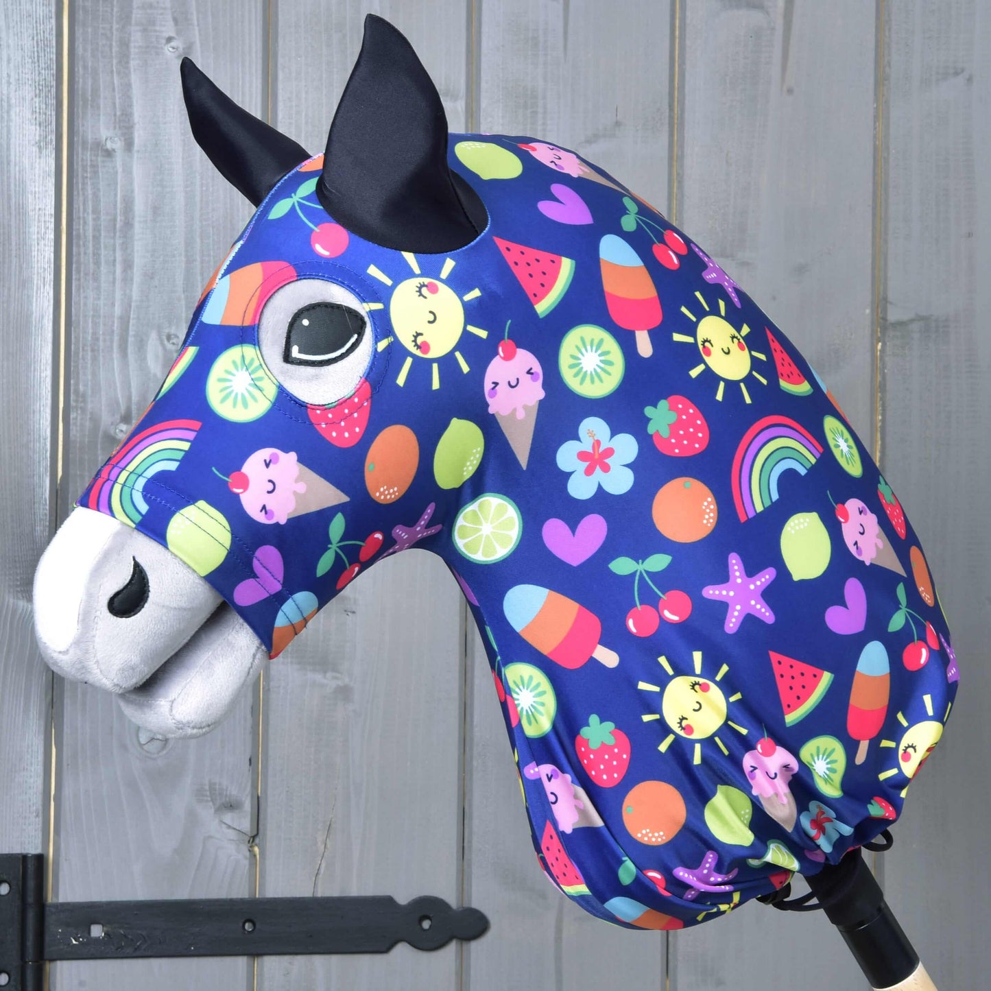Látková hlava Hobby Horse s černýma ušima, velkýma očima a modrým ochranným oblekem Hobby Horse Happiness od Hobby Horse LarDen se slunci, duhami, srdíčky, ovocem a zmrzlinou, na dřevěném pozadí.