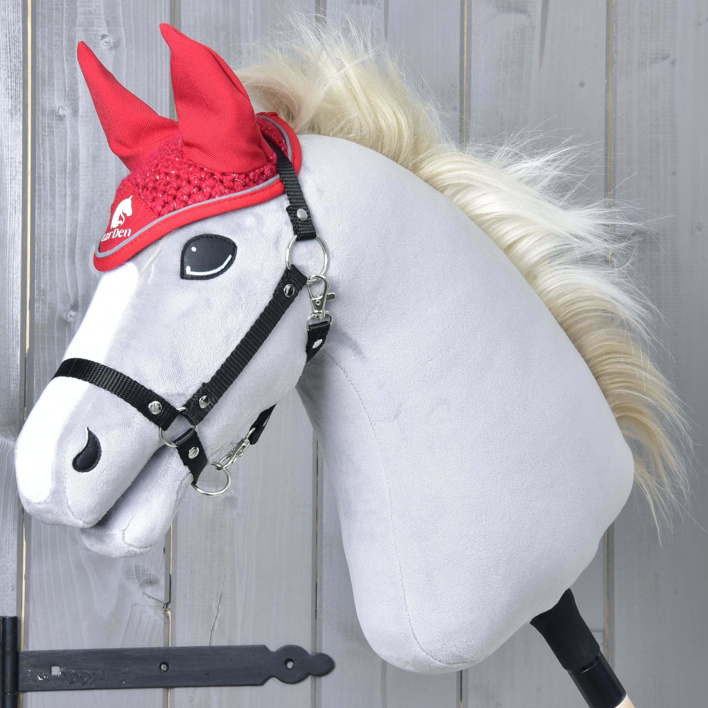 Plyšový Hobby Horse od Hobby Horse LarDen s bílým obličejem, černou uzdečkou a blonďatou hřívou předvádí čabraku Čabraka háčkovaná Red sport před světlou dřevěnou stěnou.