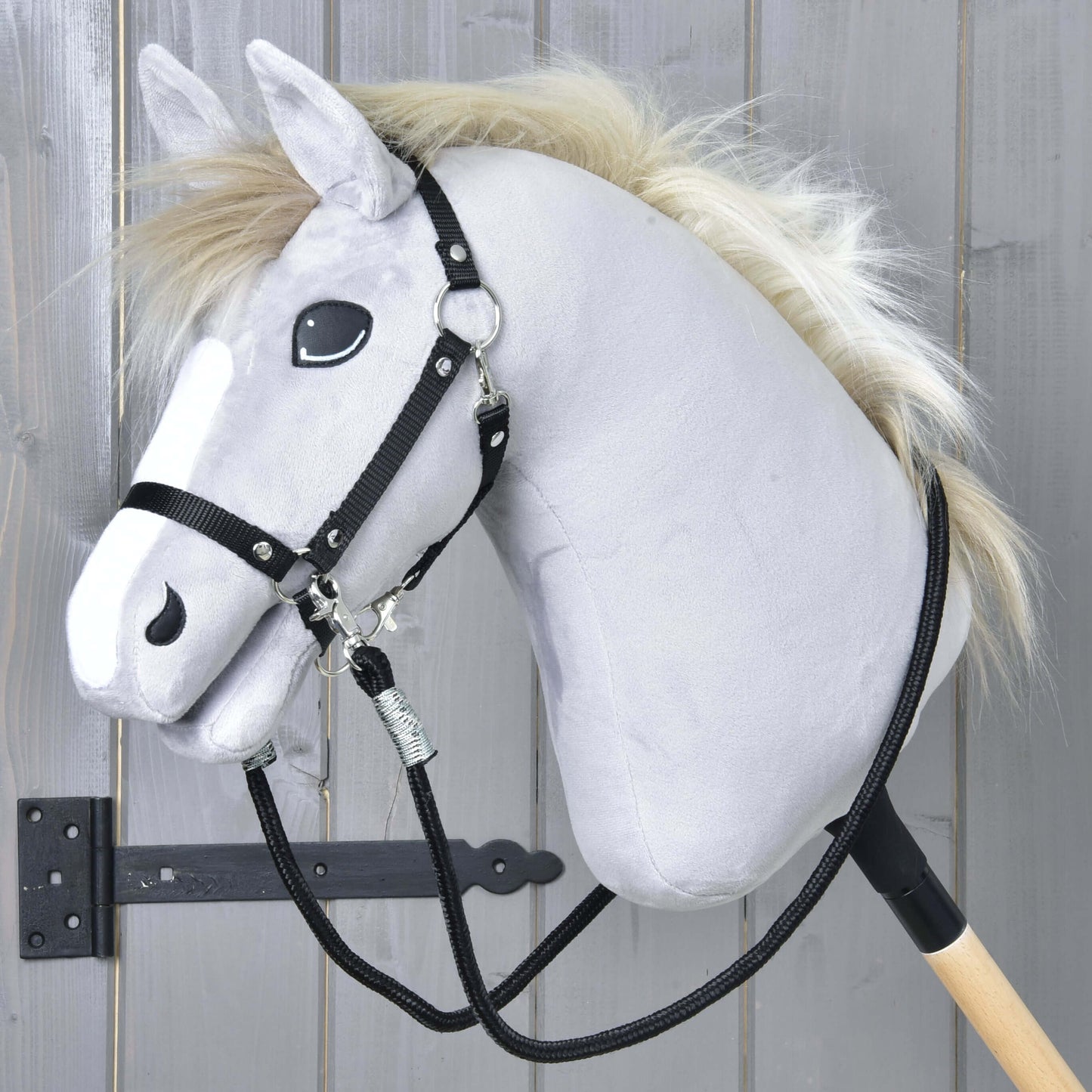 Hobby Horse - Otěže černé od Hobby Horse LarDen, s plyšovou šedou hlavou, černou uzdečkou, paracord otěžemi a bílou hřívou na dřevěné tyči, před šedými dřevěnými panely s viditelným černým kovovým pantem.