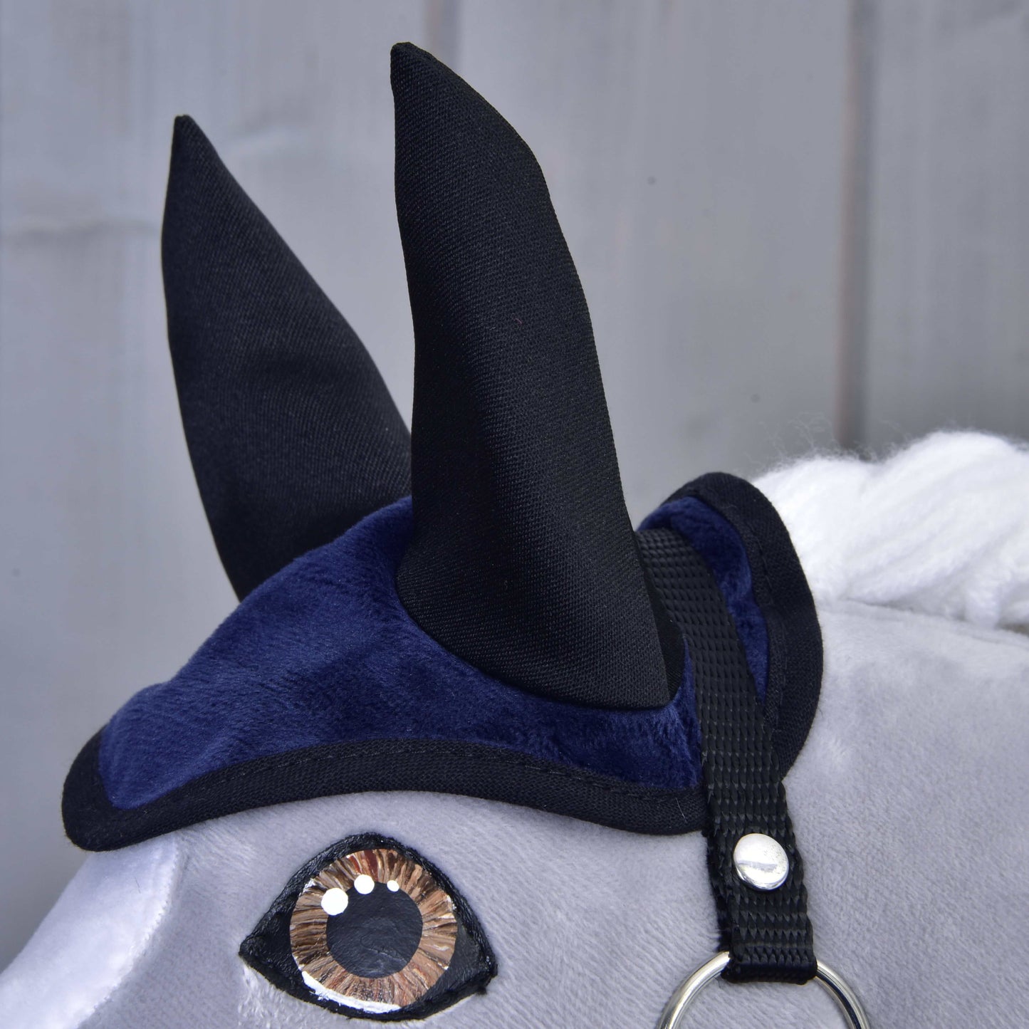 Detail hlavy plyšového hračkového koně s hnědýma očima, bílou hřívou a tmavě modro-černou čabrakou ze Set Dark Blue (čabraka + deka) od Hobby Horse LarDen, zobrazený na rozmazaném šedém pozadí.