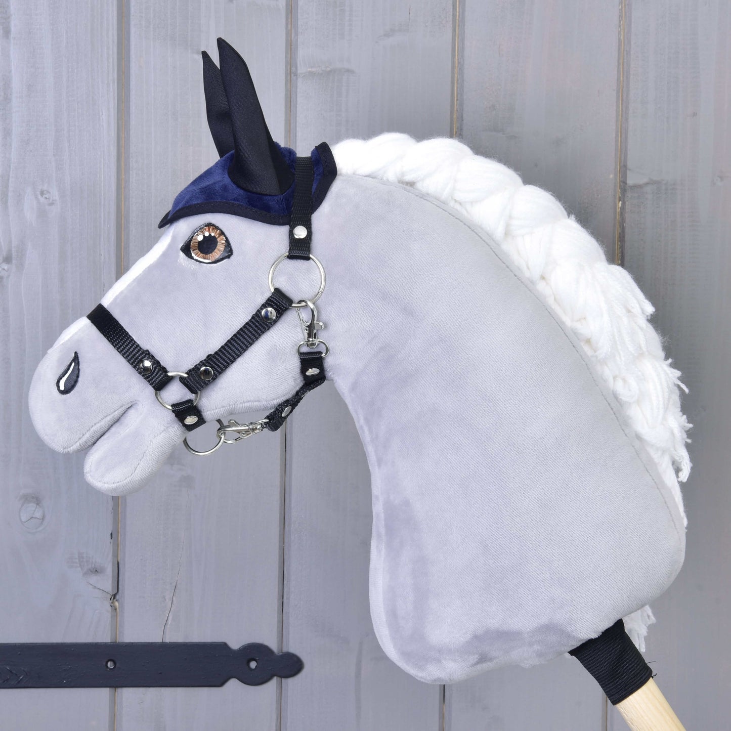 Plyšový Hobby Horse LarDen s bílou hřívou, černou uzdou a Set Dark Blue (čabraka + deka), nasazený na dřevěné tyči před světle šedou dřevěnou stěnou.