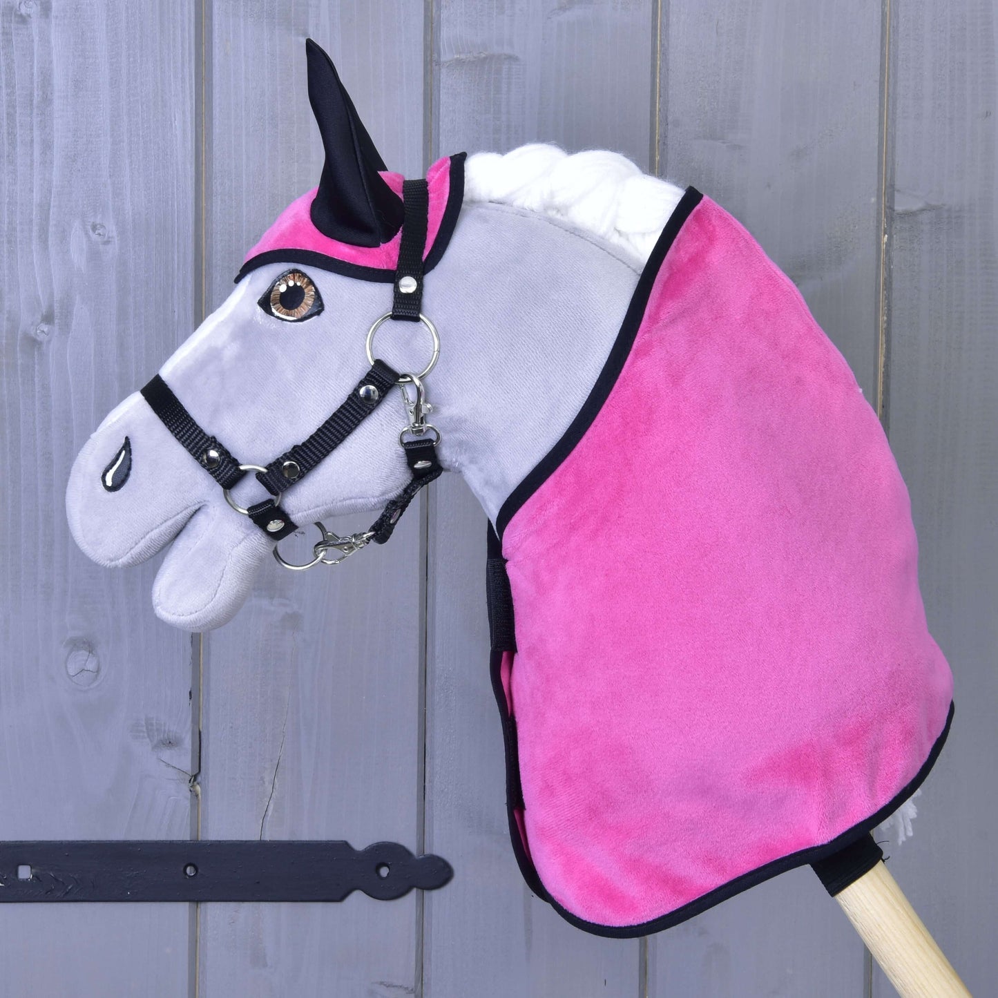 Šedá hračka koně na tyči od Hobby Horse LarDen se setem Set Hot Pink (čabraka + deka) a černou uzdečkou, umístěná před dřevěným plotem.