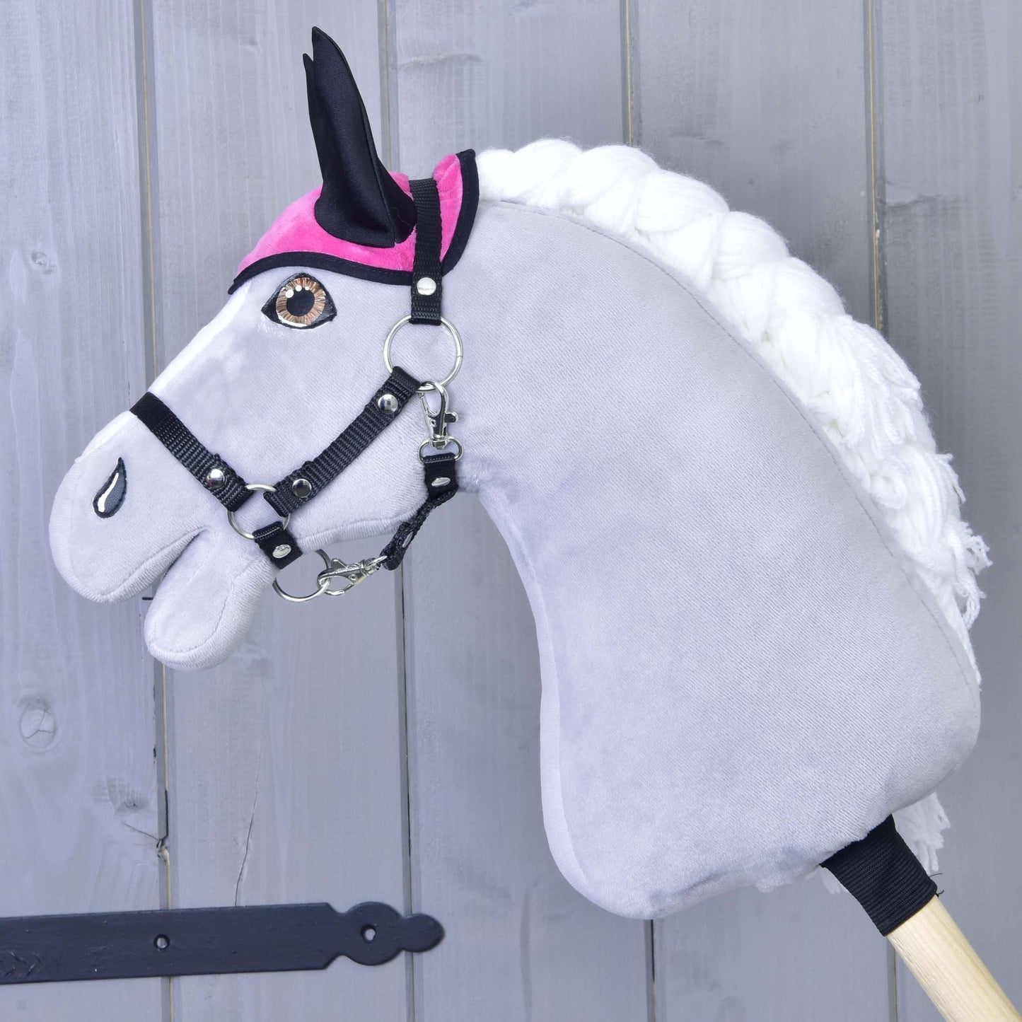 Set Hot Pink (čabraka + deka) od Hobby Horse LarDen obsahuje čabraku a deku pro váš plyšový šedý Hobby Horse s bílou hřívou, černou uzdečkou a růžovými doplňky, upevněný na dřevěné tyči před světle šedou dřevěnou stěnou.