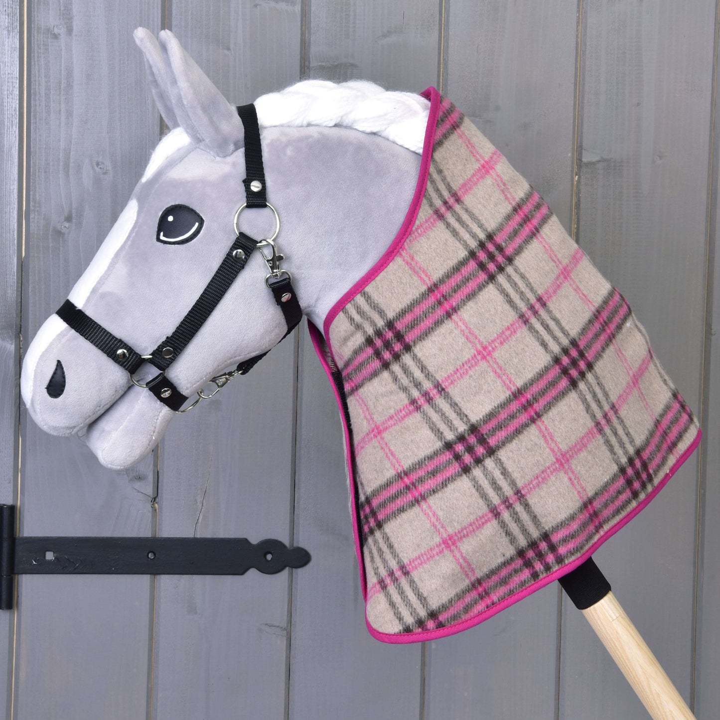 Šedý plyšový Hobby Horse LarDen s černým postrojem a kostkovaným dekovým setem Set Cube Pink přehozeným přes krk, na dřevěném pozadí.